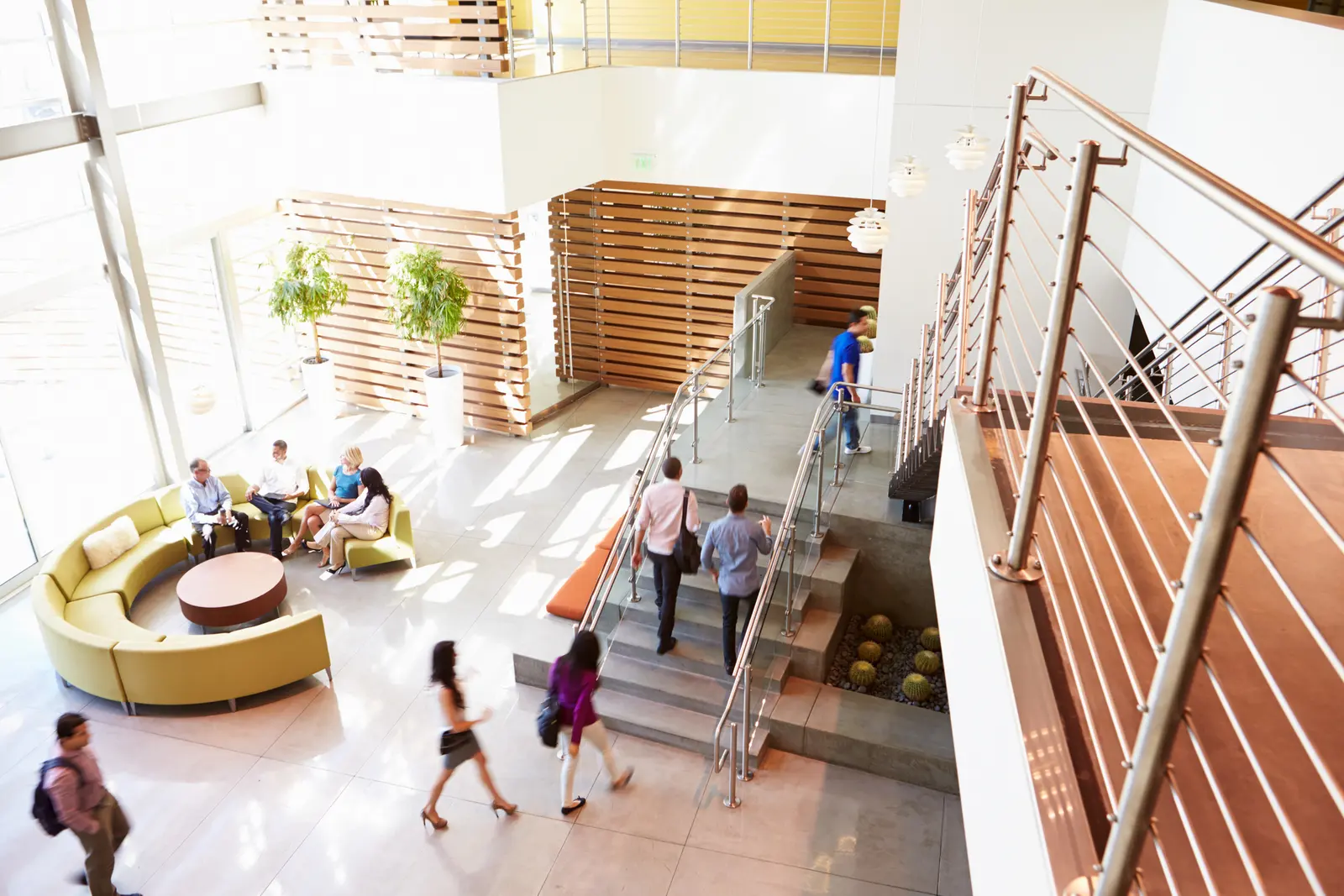 People crossing a sunlit office lobby with a lounge and open stair